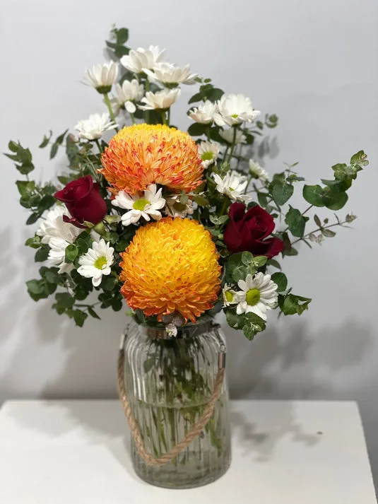 Orange chrysanthemum, white daisies, red roses and eucalyptus in a twine-wrapped glass jar